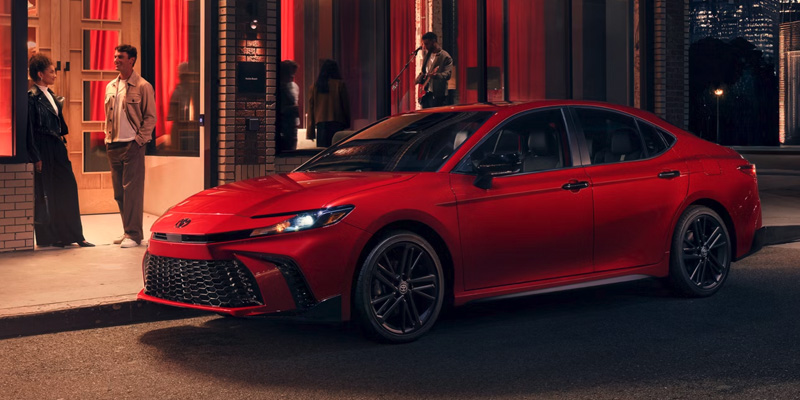 A red Toyota Camry parked on a city street at night, with people standing nearby and a vibrant backdrop.