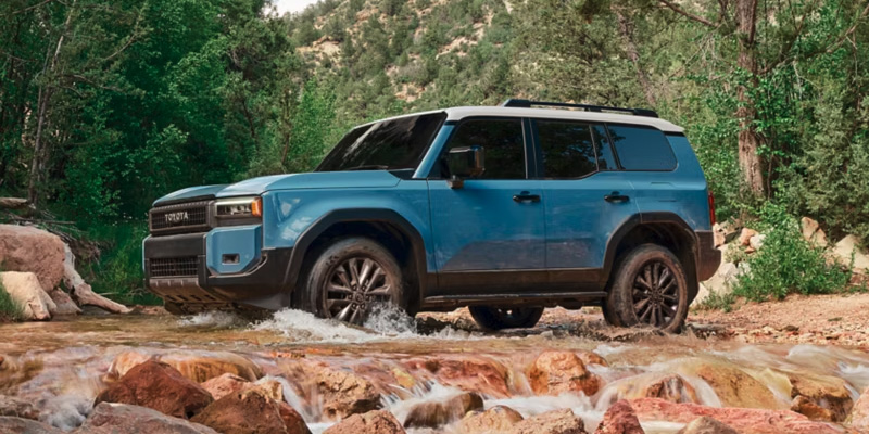 Blue Toyota SUV driving through a rocky stream in a forest landscape.
